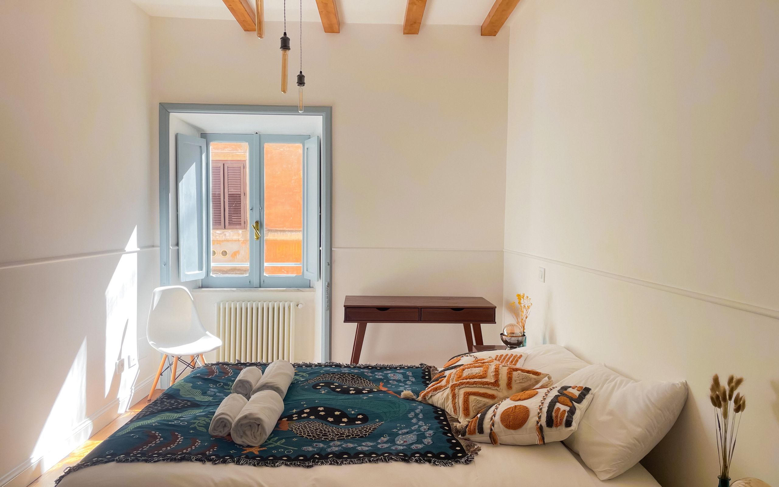Bedroom with a queen bed under a patterned teal throw, rolled towels laid out, a shuttered window open onto an ochre building, and a wooden bench along the wall