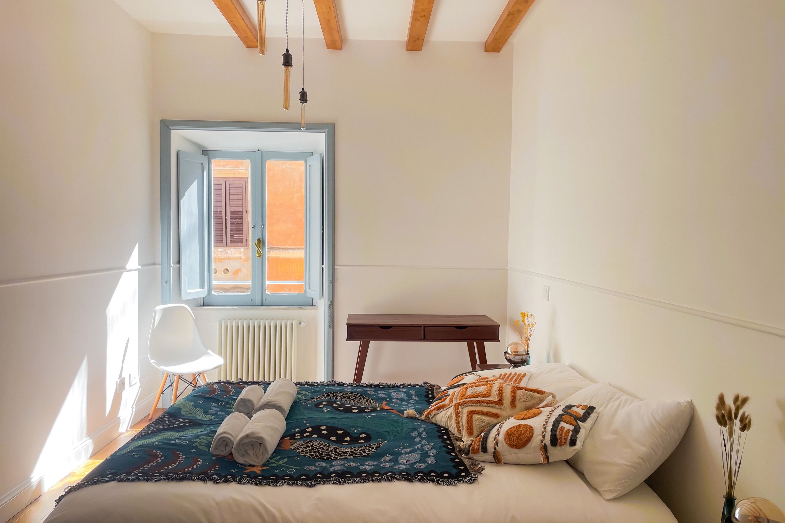 Bedroom with a queen bed under a patterned teal throw, rolled towels laid out, a shuttered window open onto an ochre building, and a wooden bench along the wall