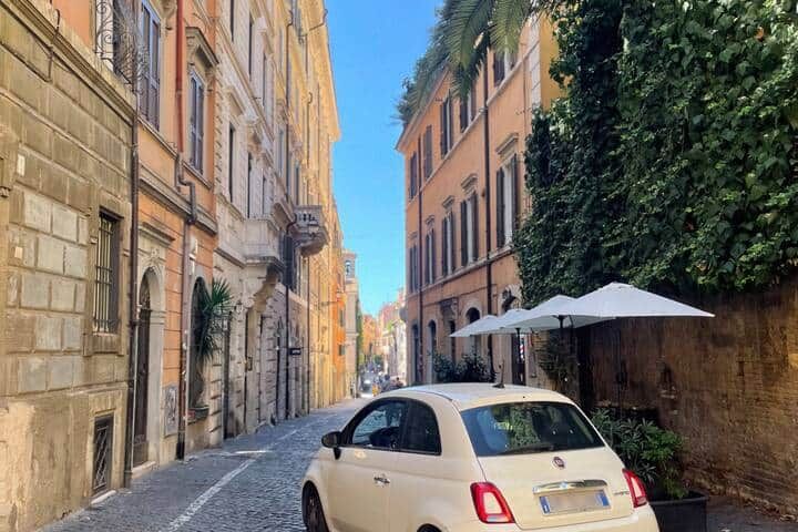 A cream Fiat 500 parked on a cobbled Monti lane beside a palm tree and a restaurant parasol, tall ochre buildings on either side