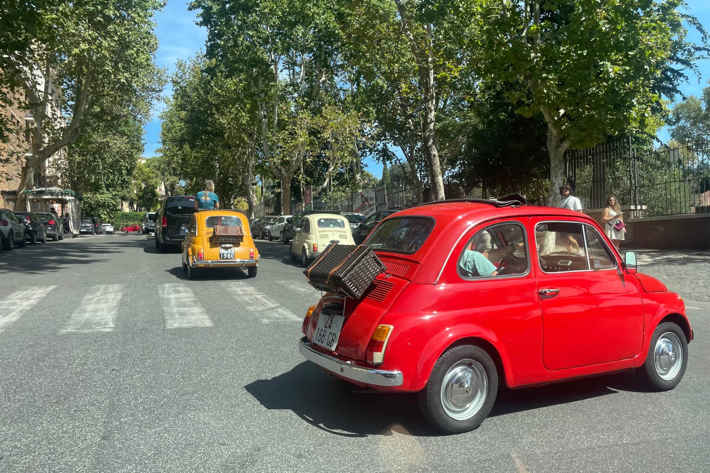 Three vintage Fiat 500s in red, yellow and cream driving down a plane-tree-lined Roman avenue, the red one carrying a wicker case on its rear rack