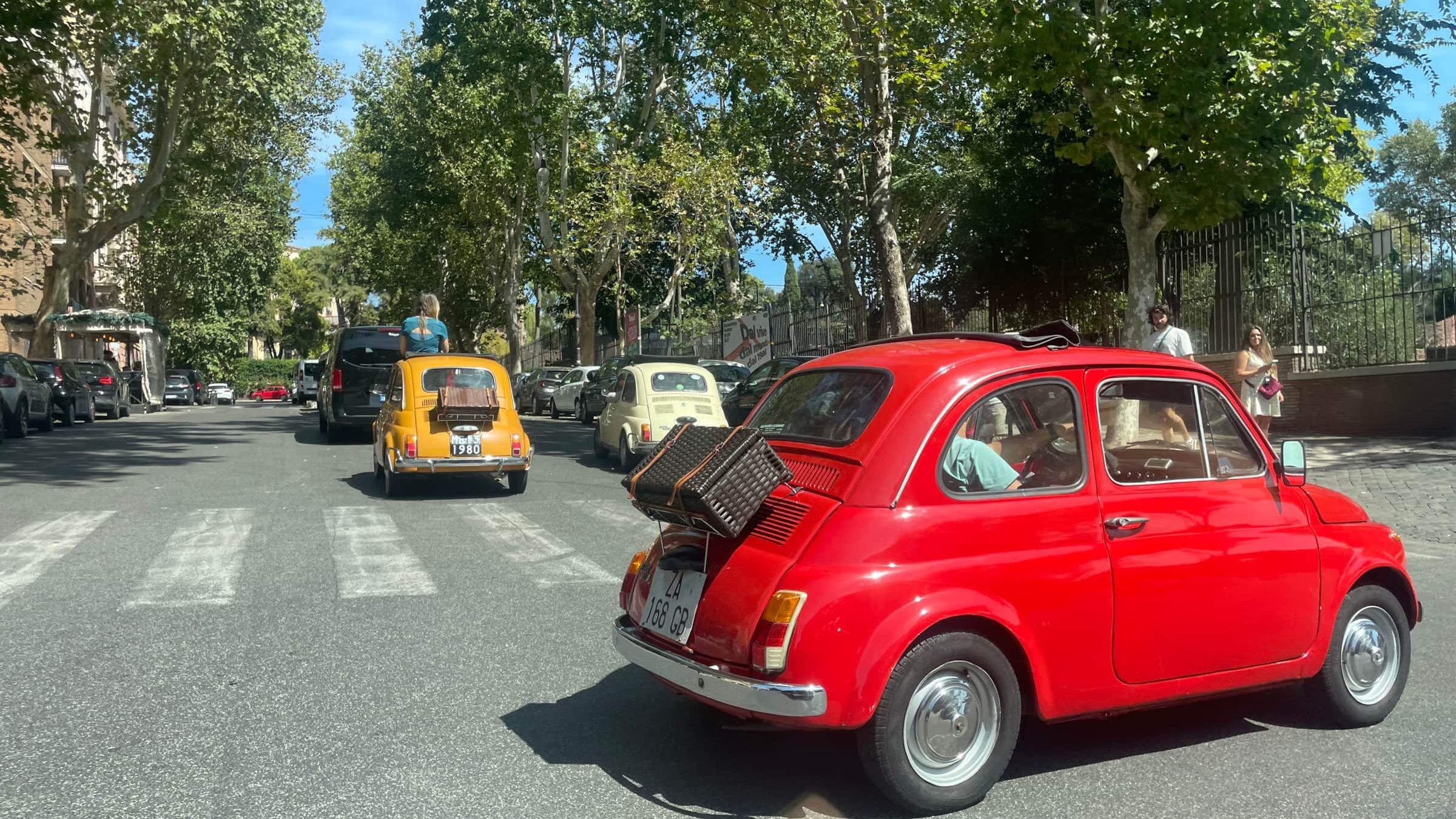 Three vintage Fiat 500s in red, yellow and cream driving down a plane-tree-lined Roman avenue, the red one carrying a wicker case on its rear rack