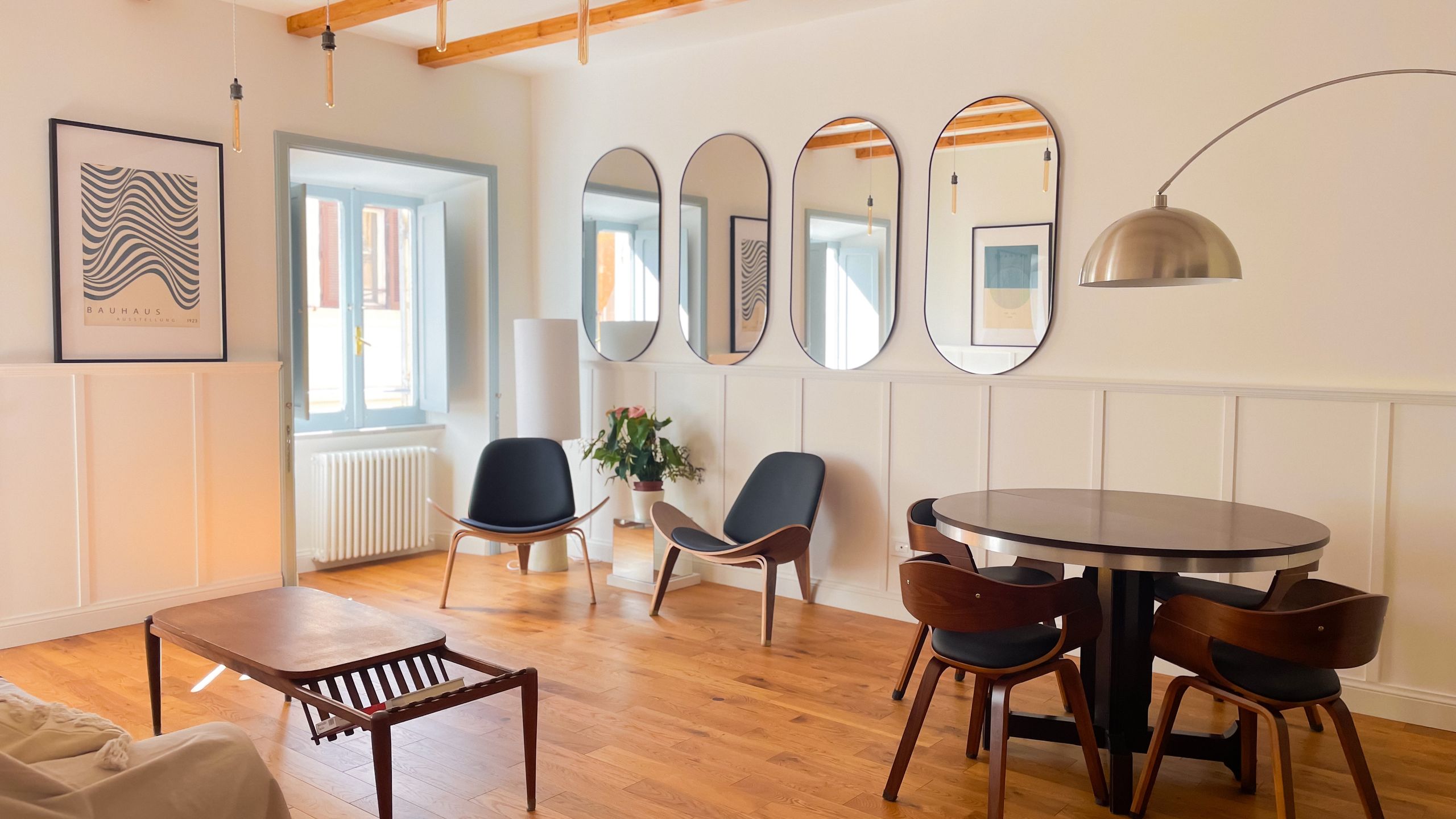 Living room with a round wooden dining table, four curved-back chairs, a row of oval mirrors along the wall and an arc floor lamp, under exposed timber beams