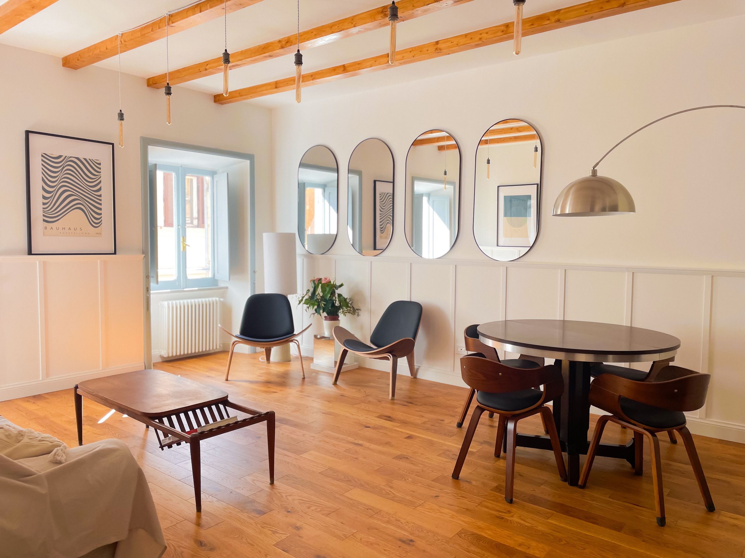 Living room with a round wooden dining table, four curved-back chairs, a row of oval mirrors along the wall and an arc floor lamp, under exposed timber beams