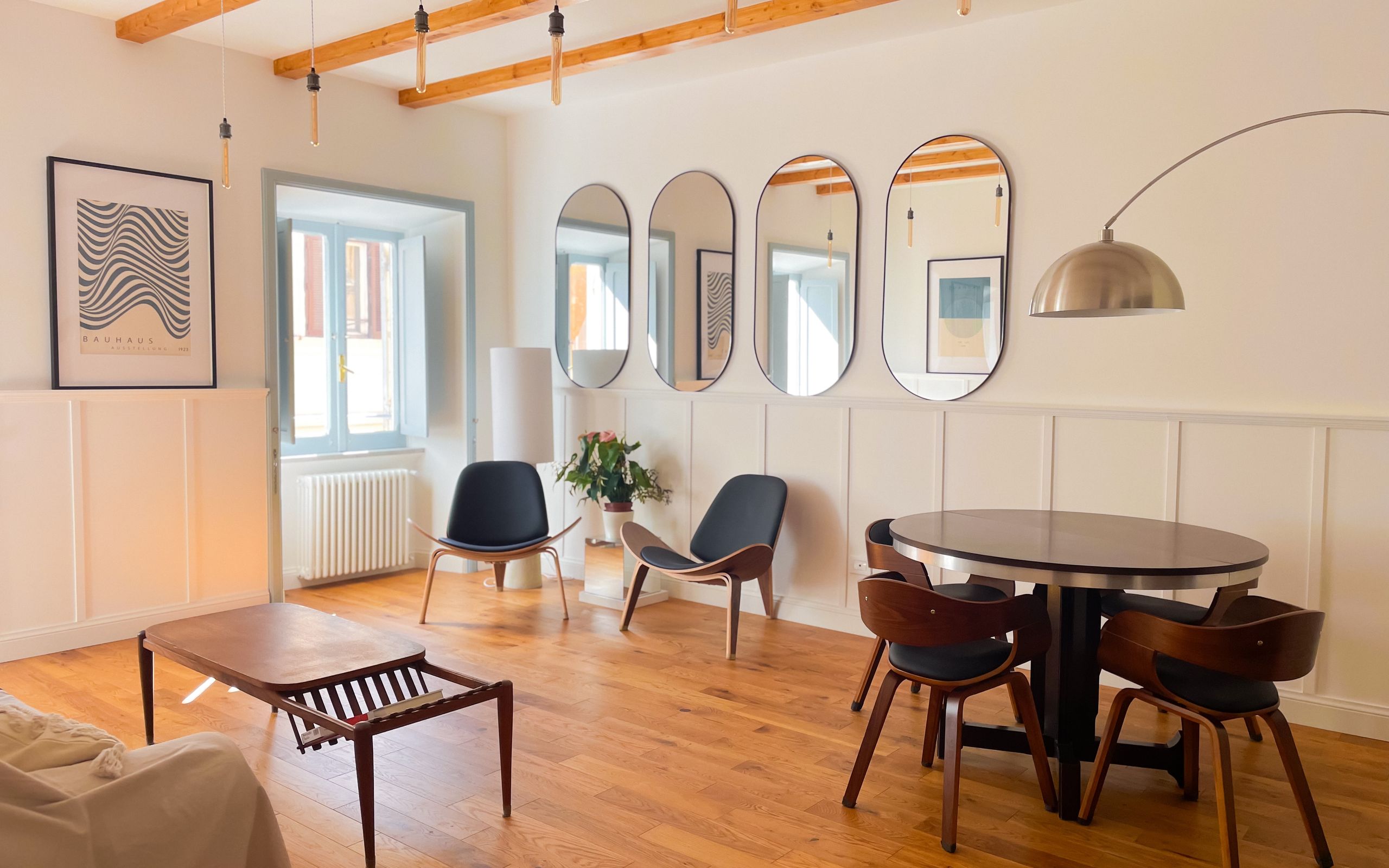 Living room with a round wooden dining table, four curved-back chairs, a row of oval mirrors along the wall and an arc floor lamp, under exposed timber beams
