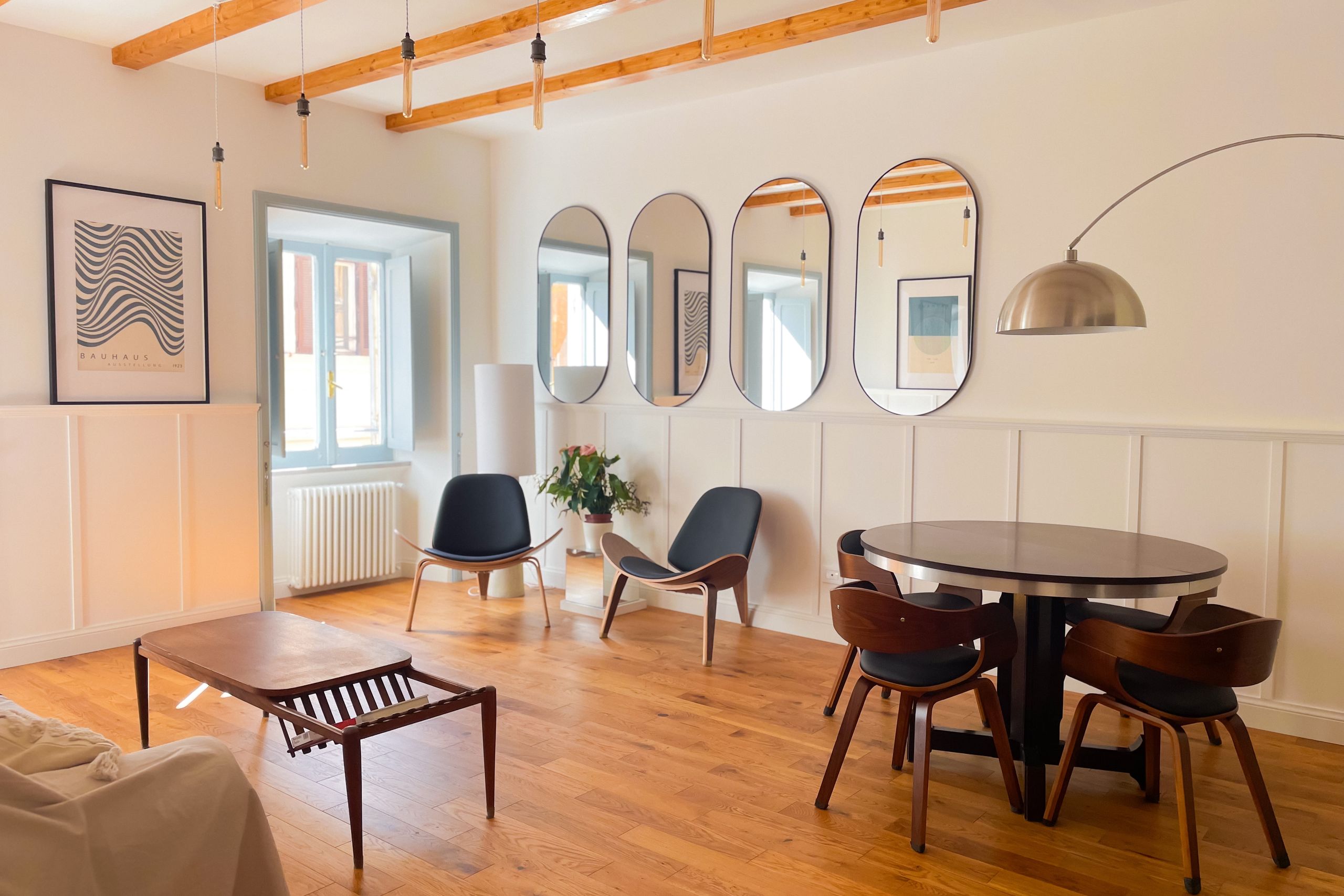 Living room with a round wooden dining table, four curved-back chairs, a row of oval mirrors along the wall and an arc floor lamp, under exposed timber beams