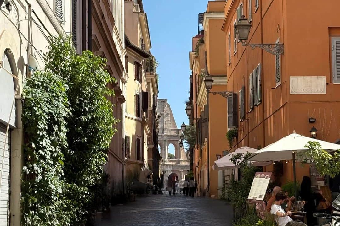 A cobbled lane between ochre and cream buildings in Monti, with the arches of the Colosseum framed at the far end and people at café tables in the foreground