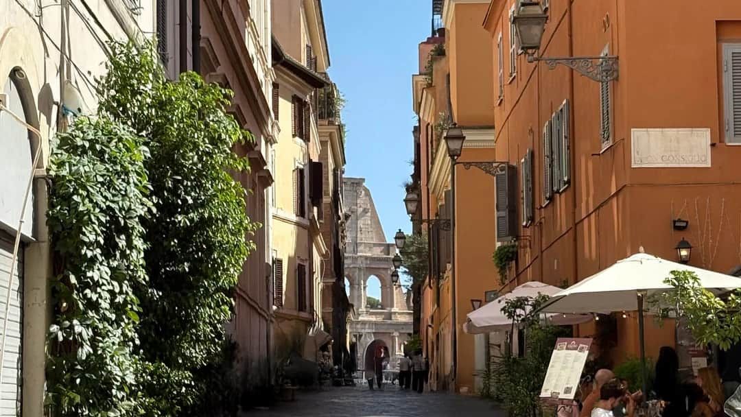 A cobbled lane between ochre and cream buildings in Monti, with the arches of the Colosseum framed at the far end and people at café tables in the foreground