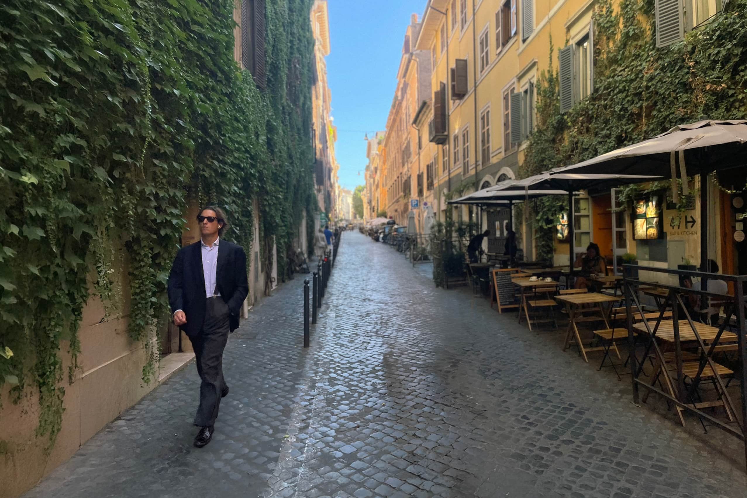 A cobbled Monti lane with an ivy-covered wall on one side and café tables under parasols on the other, a man in a suit walking towards the camera