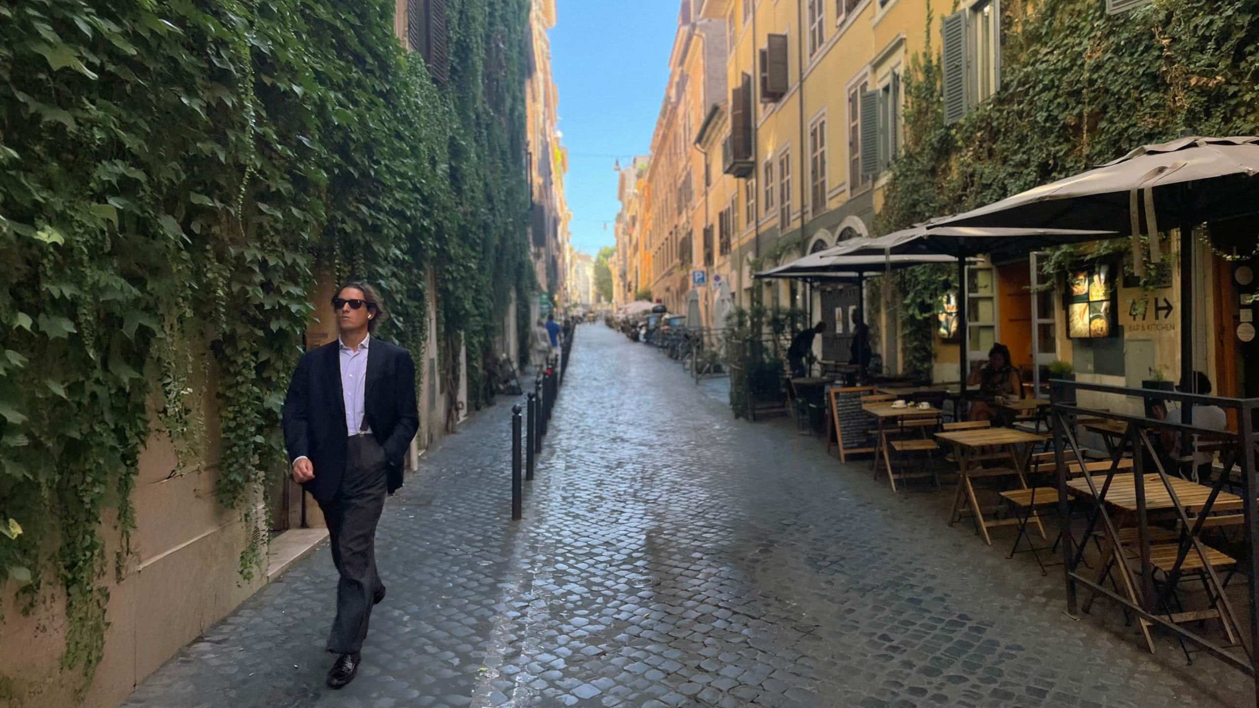 A cobbled Monti lane with an ivy-covered wall on one side and café tables under parasols on the other, a man in a suit walking towards the camera
