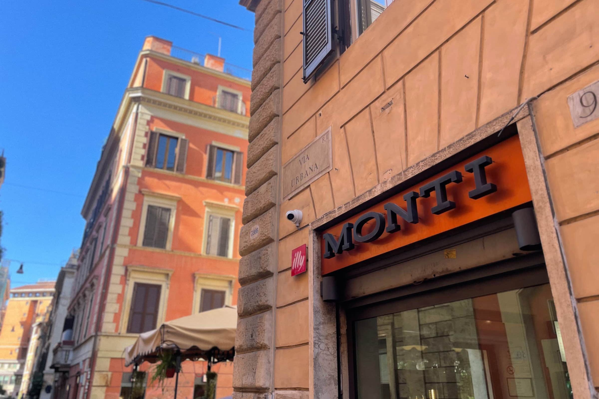 The marble street plaque reading Via Urbana on a terracotta building, beside a shopfront with MONTI in black letters, under a deep blue sky