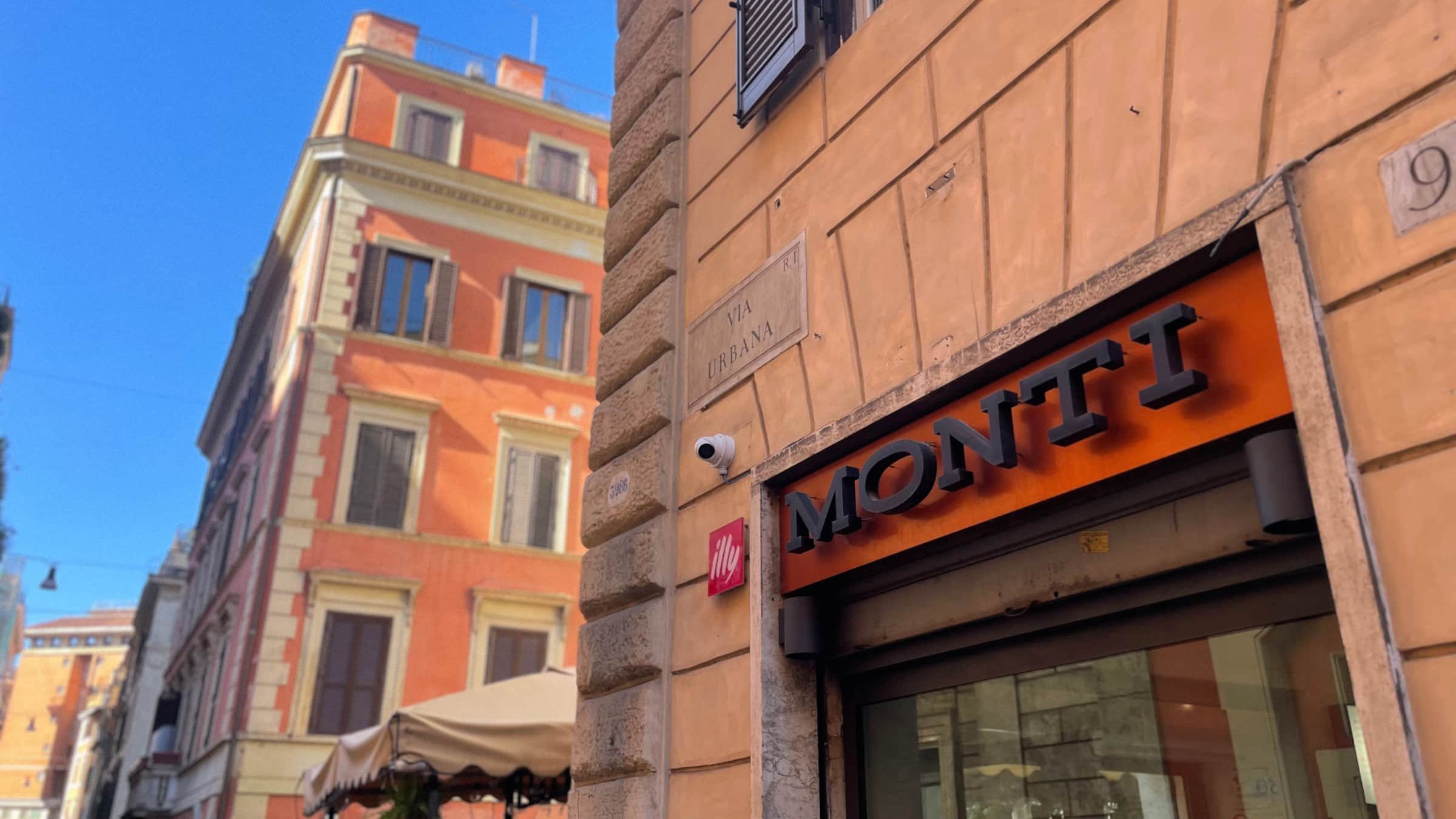 The marble street plaque reading Via Urbana on a terracotta building, beside a shopfront with MONTI in black letters, under a deep blue sky