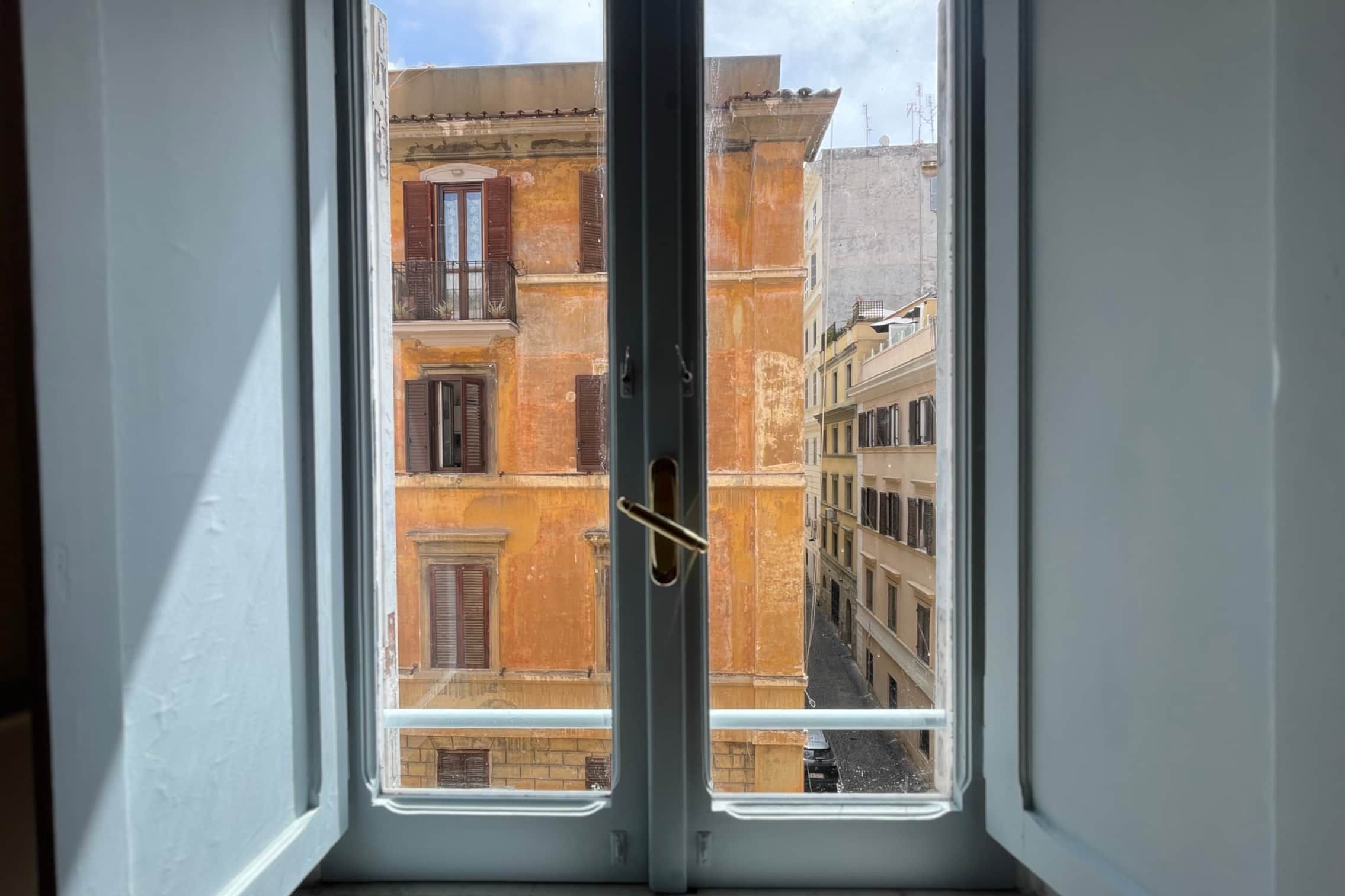 The view out of an open casement window in the apartment, across a narrow street to a weathered ochre façade with green shutters