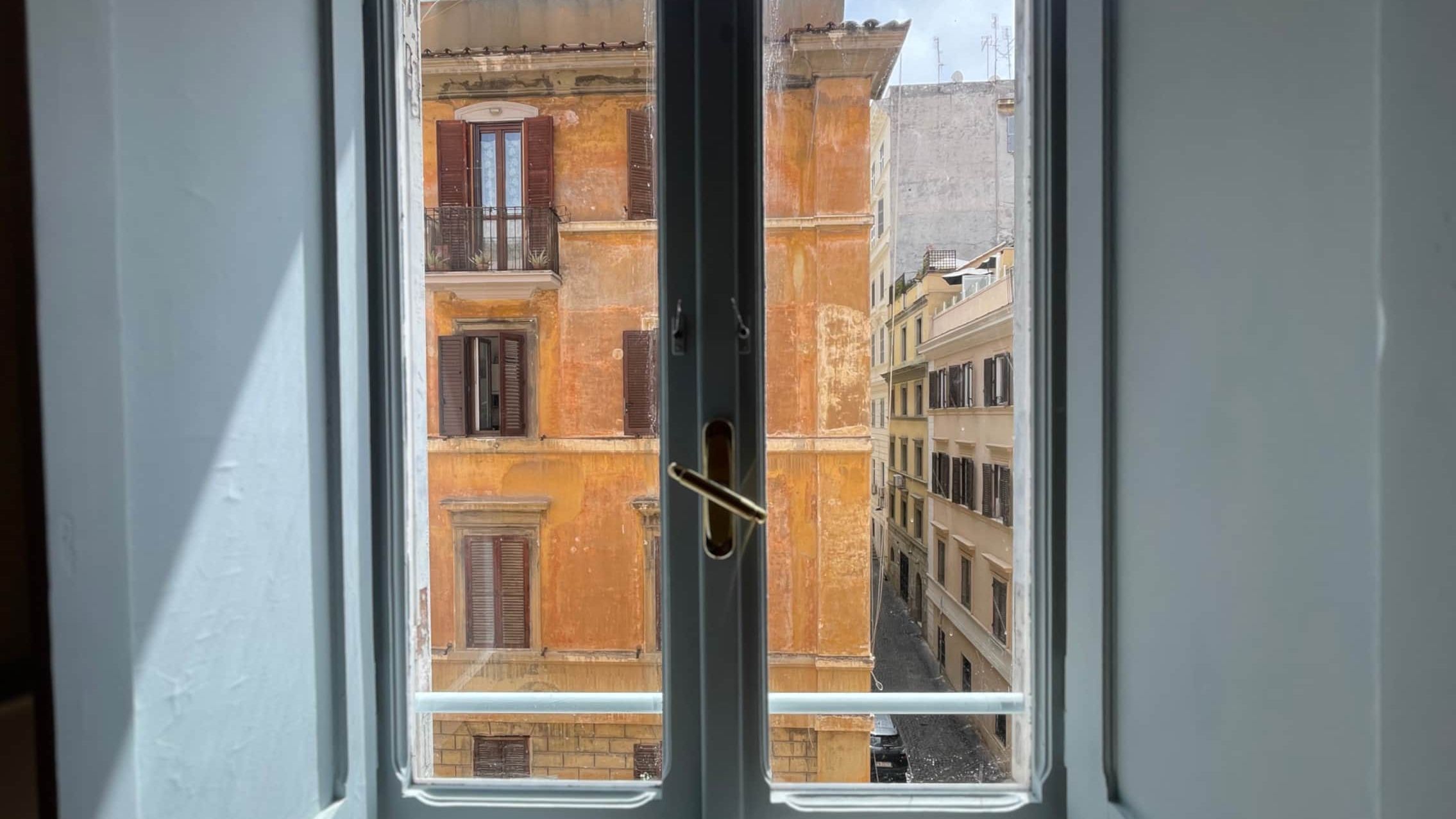 The view out of an open casement window in the apartment, across a narrow street to a weathered ochre façade with green shutters